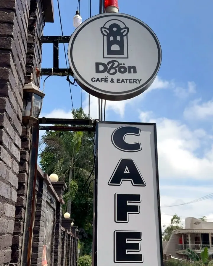 Papan bulat berlogo DBon Cafe & Eatery dan papan vertikal bertuliskan CAFE di dinding bata hitam
