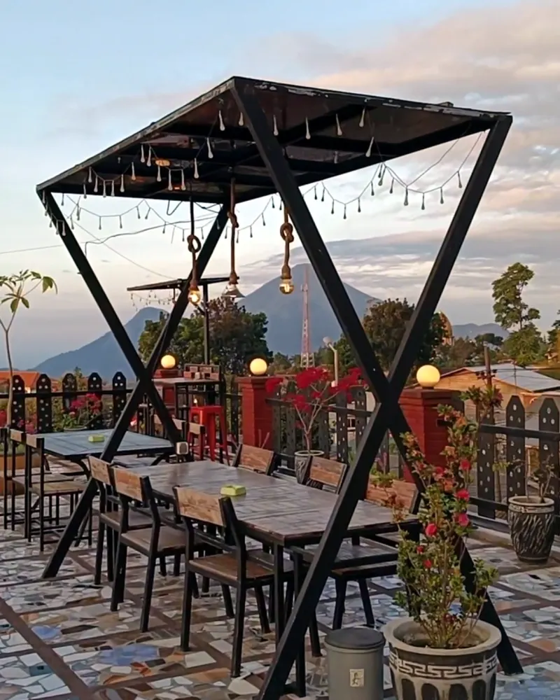 Area duduk rooftop DBon dengan rangka besi dan lampu gantung, berlatar langit sore dan pegunungan