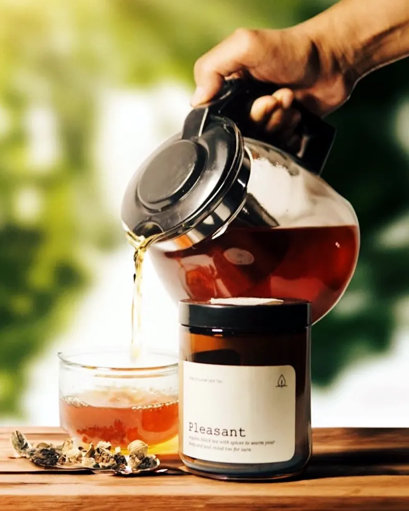 Teh artisan dituang dari teko kaca ke gelas, di samping toples teh Pleasant di meja kayu